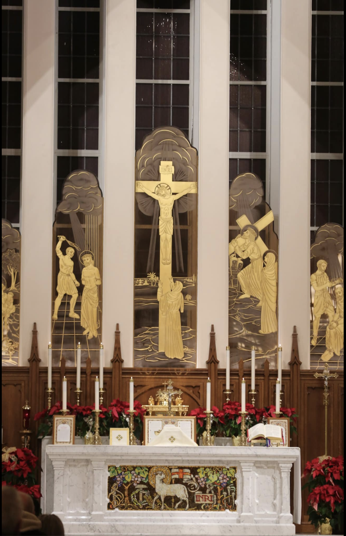 White high altar with a Lamb of God front panel, candlesticks and poinsettias, below gold relief panels of the Crucifixion